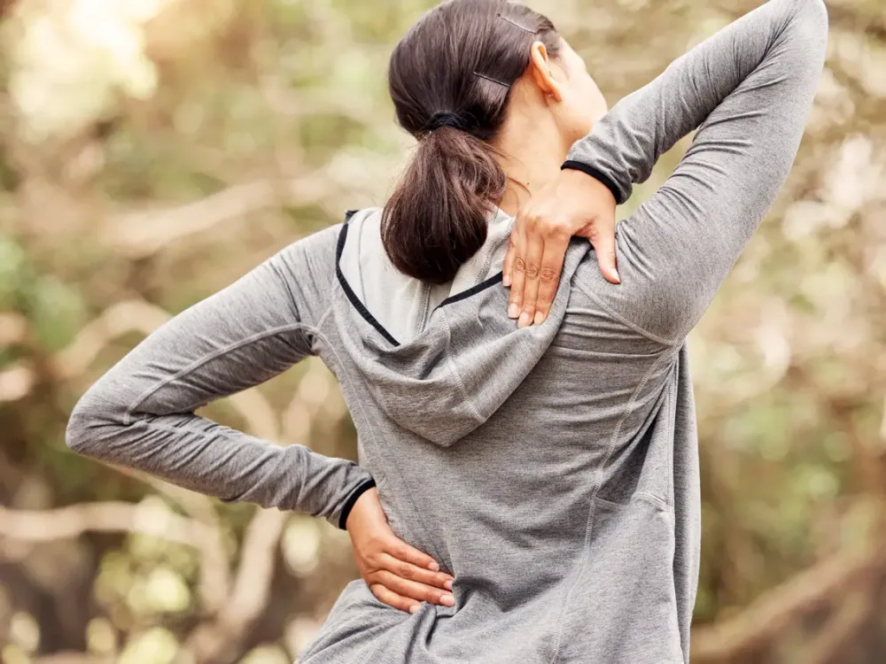 A woman outdoors reaches behind her back and shoulder in discomfort, showing the kind of lower back pain that can radiate down the leg as sciatica.