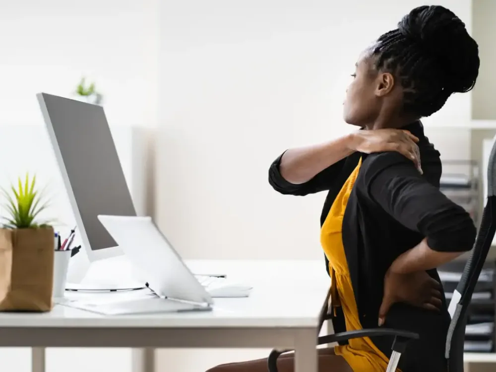 A woman at her office desk holds her neck and lower back, illustrating the persistent day-to-day pain that defines a chronic pain condition.