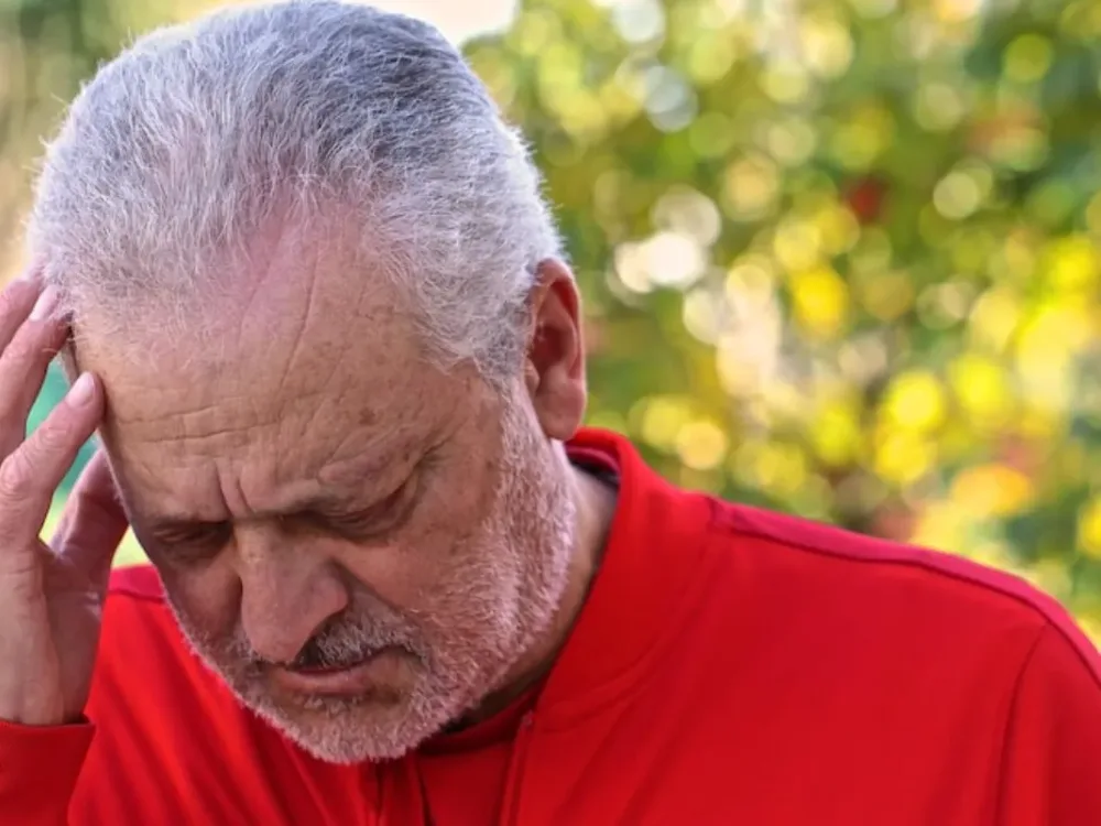 An older man holds his temple outdoors, wincing from head pain that often originates in the neck and upper back.