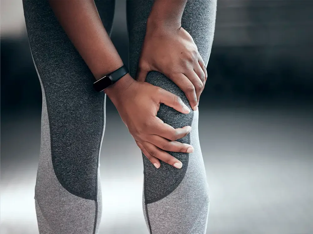 A woman in athletic leggings grips her knee with both hands, showing the hip-and-knee compensation pattern we assess in treatment.
