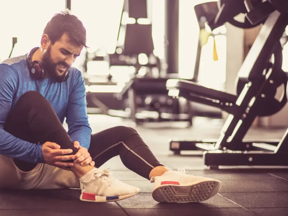 A runner sits on the gym floor holding his shin and ankle after training, showing the kind of running injury we build a comeback plan around.