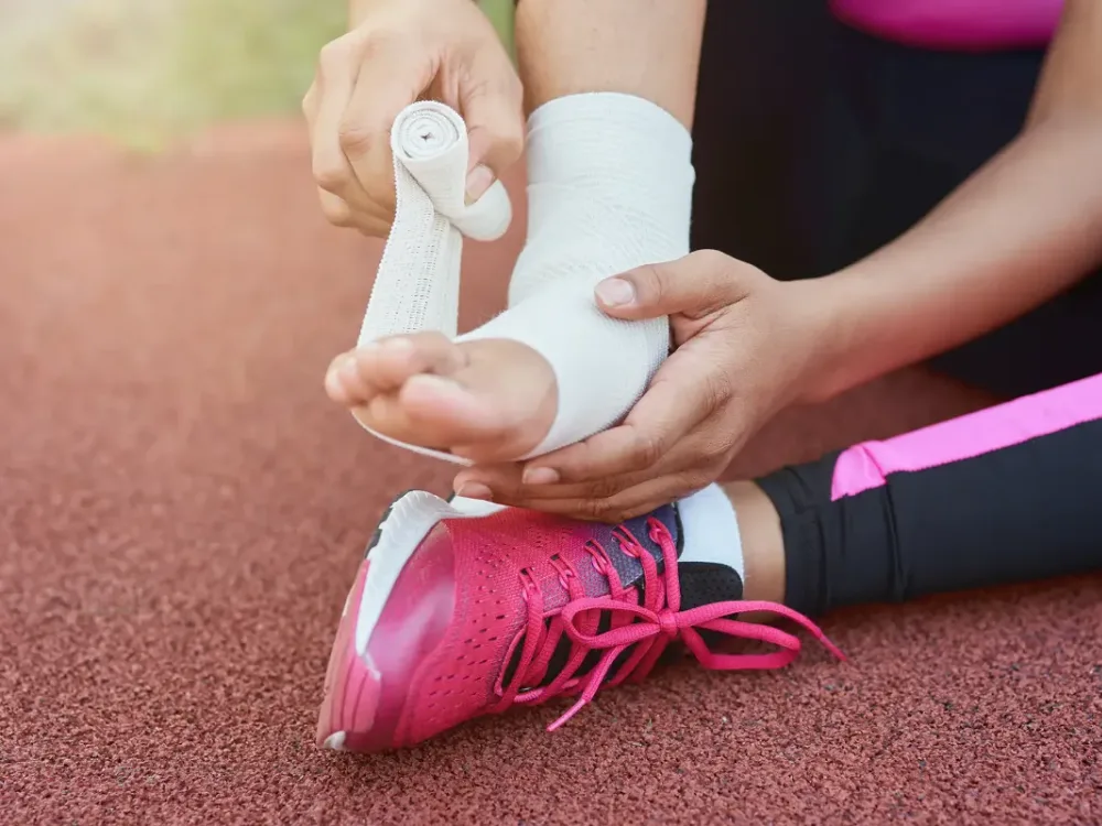 An athlete wraps her ankle with athletic tape on the track, illustrating the sprains and strains we rebuild before clearing a return to play.