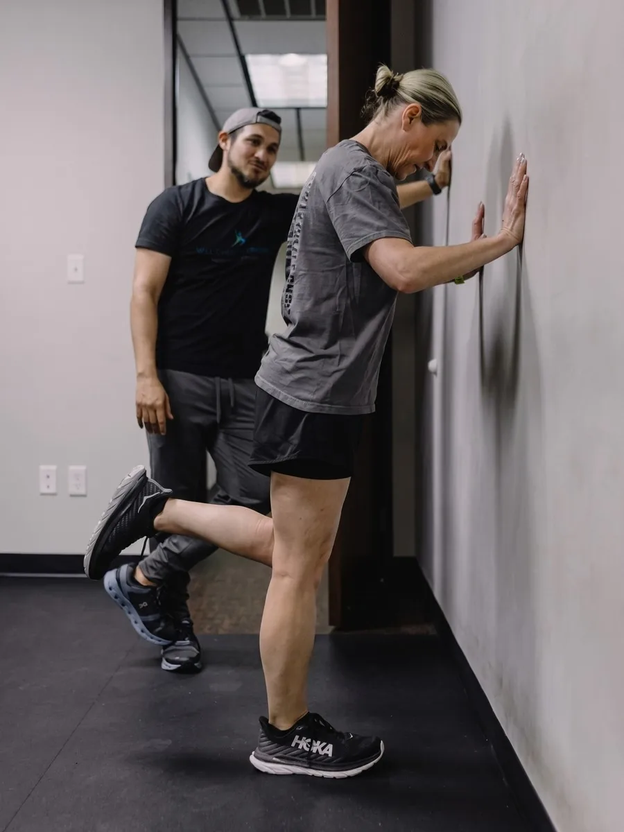 A trainer helps a client hold a supported single-leg balance stretch against a wall.