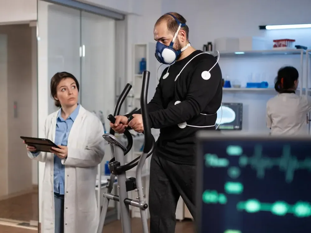 A runner wearing motion-capture sensors and a metabolic mask runs on an instrumented treadmill while a clinician monitors live biomechanics data on a screen.