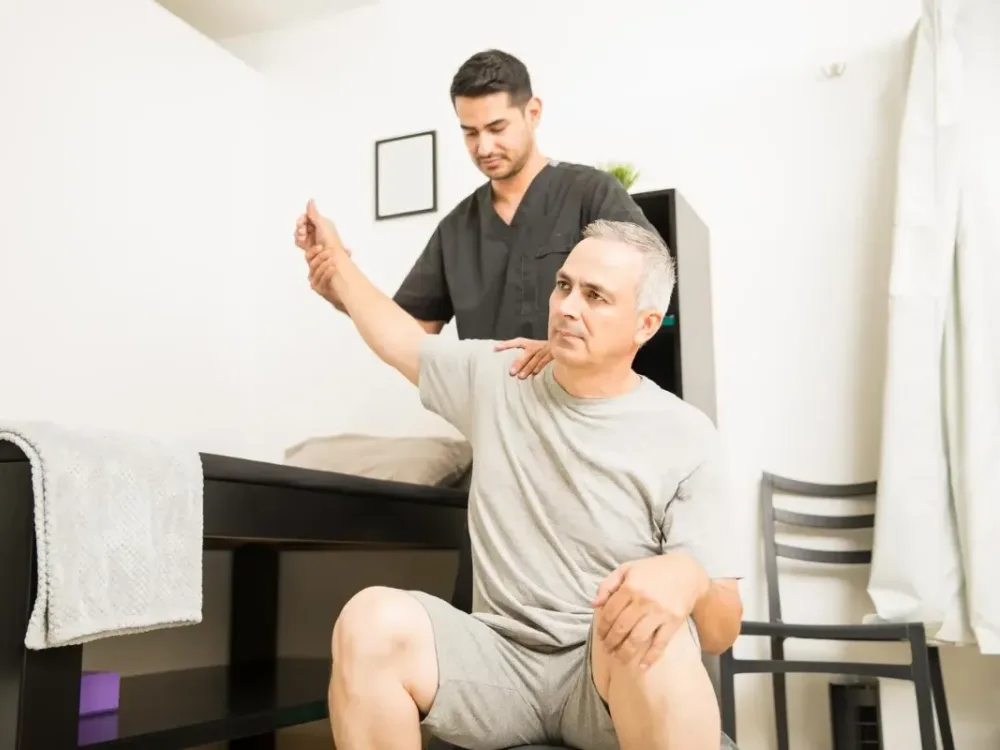 A clinician guides a seated patient's arm through an overhead range-of-motion check while a hand rests on his shoulder to feel how the joint moves.