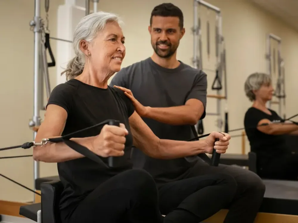 A clinician spots a patient performing a resisted seated row on reformer equipment as part of a supervised rehab exercise session.
